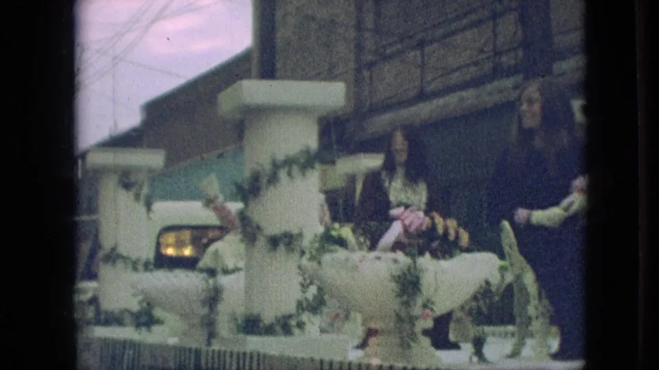 A wider 8mm frame shows two women on a decorated parade float with white pillars, green garlands, wrapped decorations, and street buildings in the background.
