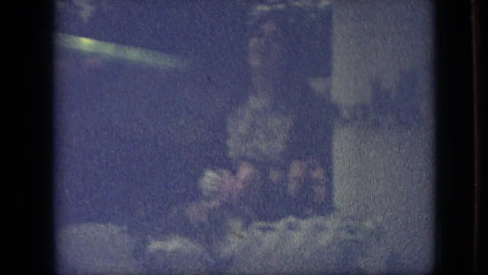 Low-light 1968 Branson, Missouri 8mm frame showing two adult figures outdoors, with one figure appearing to cradle plants or flowers near a garden or yard area.