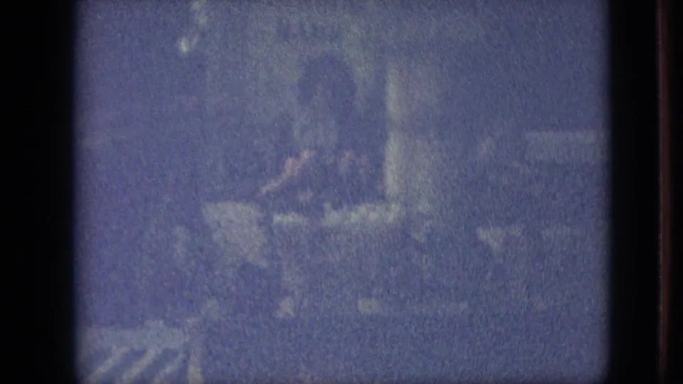 Grainy blue-tinted 1968 Branson, Missouri home-movie frame with a partially obscured adult figure near plants or flowers in a dark outdoor setting.