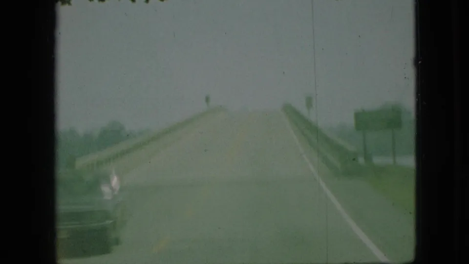 Green-tinted 1968 Missouri home-movie frame showing a hazy road ahead from a vehicle perspective, with guardrails, overcast sky, and a roadside sign shape.