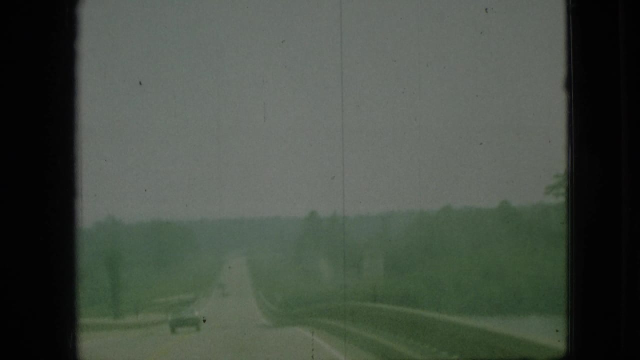1968 Branson, Missouri 8mm frame showing a forward road view from a moving vehicle, with trees, hazy sky, and a small distant car.