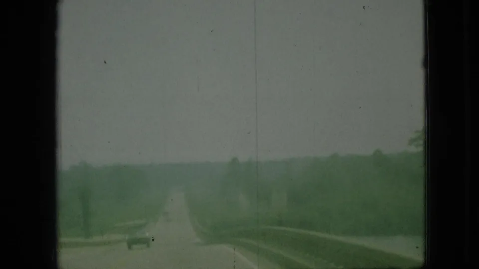 1968 Branson, Missouri 8mm frame showing a forward road view from a moving vehicle, with trees, hazy sky, and a small distant car.