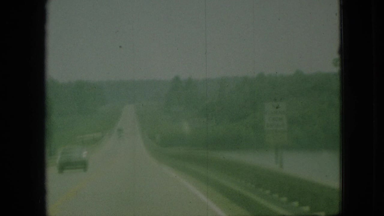 Two-lane highway beside water with a car ahead and trees in the distance.