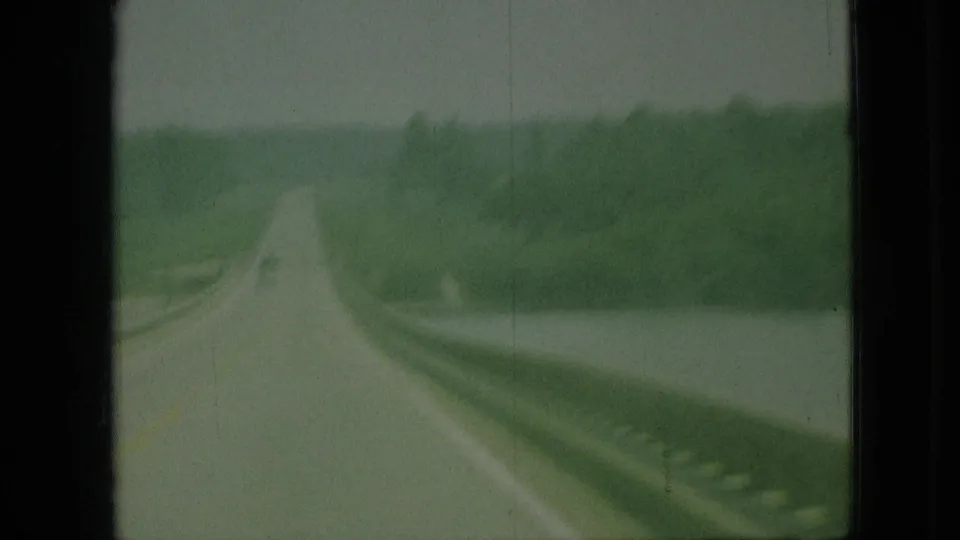 1968 Branson, Missouri 8mm frame with a curving rural road, roadside water, guardrail, and dense green tree line seen from a moving vehicle.