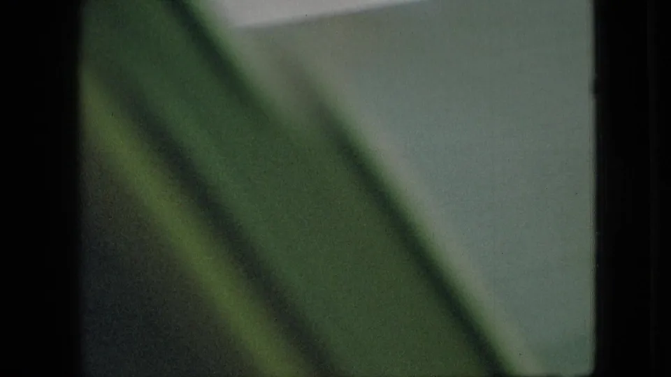 Motion-blurred 8mm frame with a green diagonal foreground shape crossing a pale exterior view, consistent with filming from a moving vehicle.