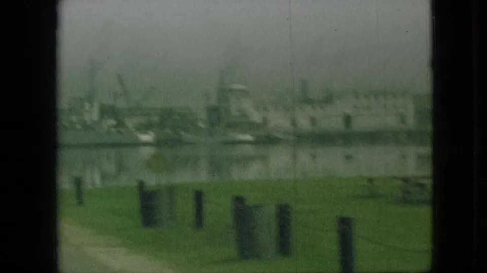 1968 Branson, Missouri home-movie frame showing a distant waterside area with grass, short posts, shoreline, and faint industrial or marina-like structures.