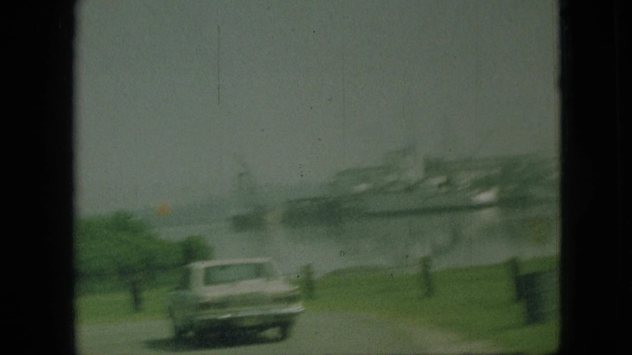 1968 Branson, Missouri home-movie frame showing roadside grass, a classic car, and a hazy waterfront with a large vessel or dock structure beyond.