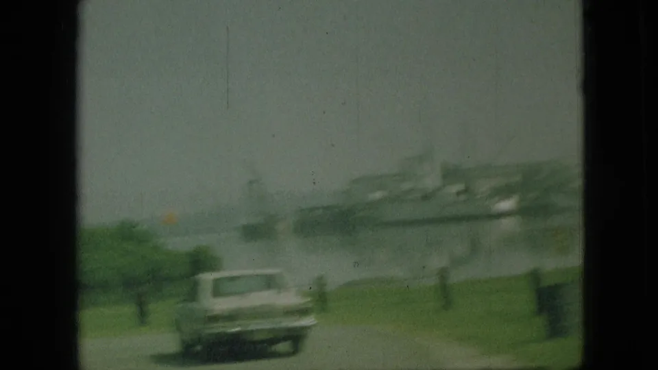1968 Branson, Missouri home-movie frame showing roadside grass, a classic car, and a hazy waterfront with a large vessel or dock structure beyond.