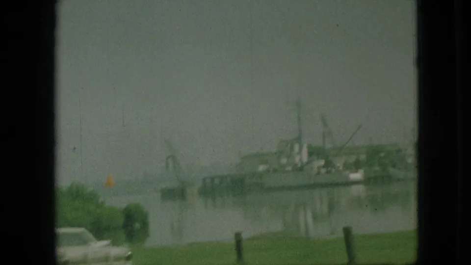 1968 Branson, Missouri frame from a moving car looking toward a waterfront with boats or dock structures, an American flag, grass, and a partial car in the foreground.