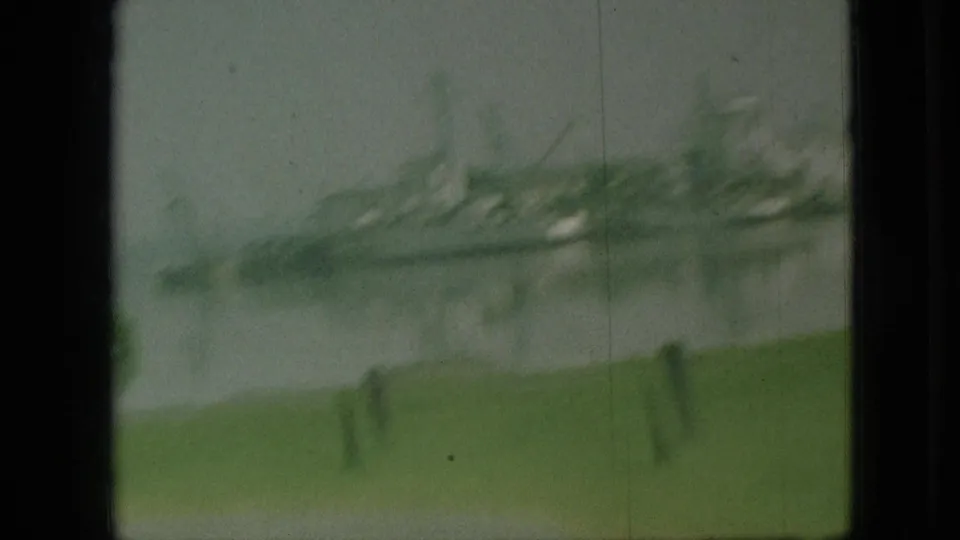 Soft 1968 Branson, Missouri home-movie frame with a blurred boat, barge, or dock structure across the water behind grass and roadside posts.