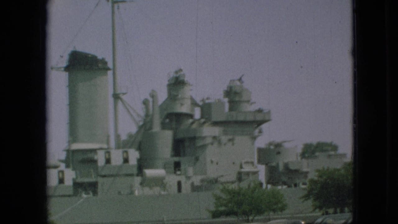 Large gray docked warship with smokestacks, superstructure, trees, and parked cars.