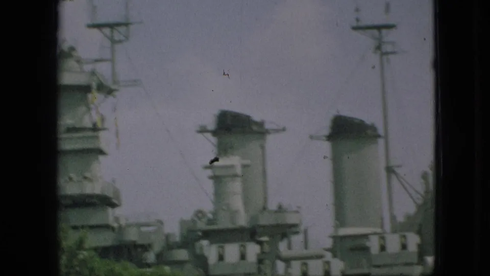 1968 Missouri-labeled home-movie frame showing the docked ship's smokestacks and upper structures with utility poles, trees, and muted sky around it.