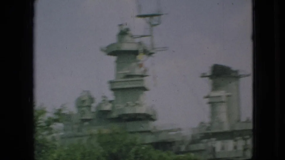 1968 Branson, Missouri home-movie frame showing a gray naval superstructure with mast and turret-like forms seen through foliage and dark vertical borders.