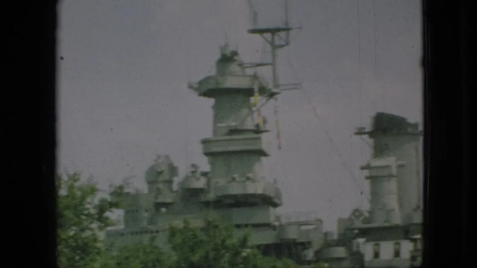 1968 Branson frame showing a closer visitor-shot view of gray battleship superstructure, antenna mast, deck details, and foreground greenery.