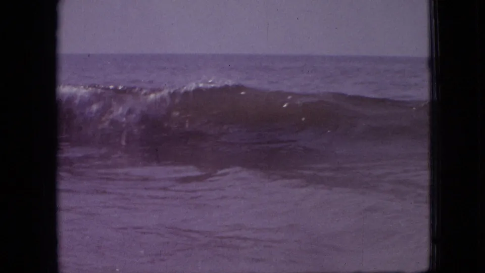 1960s 8mm home-movie frame showing a cresting wave offshore under a low horizon, with purple color shift and visible gate edges.