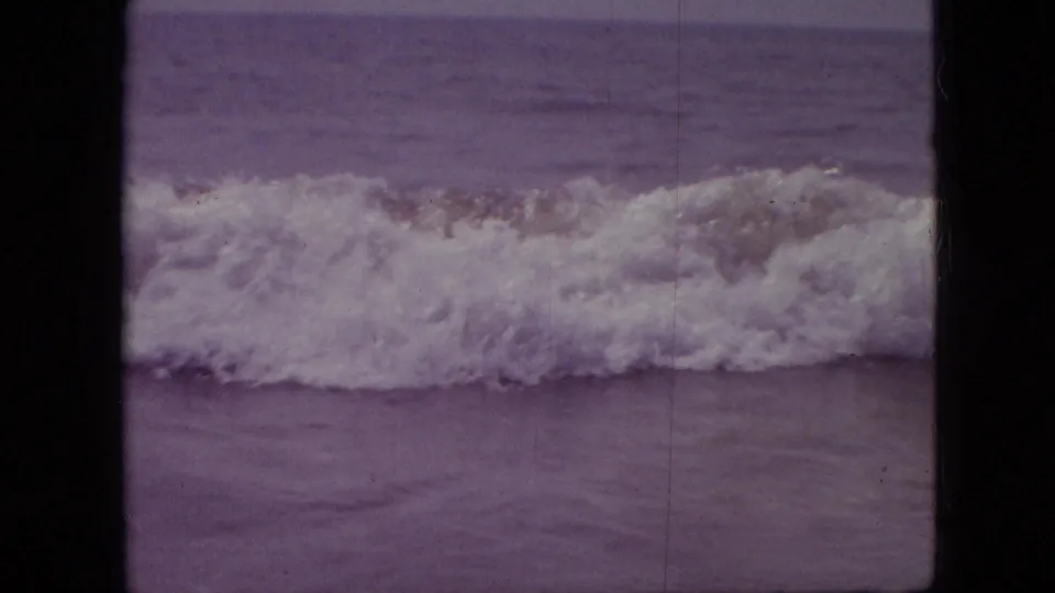 1960s 8mm home-movie frame showing white foam from a breaking wave spreading across shallow water, with scratches and aged color shift.