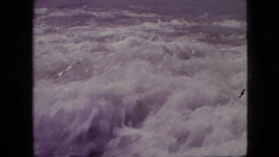 1960s 8mm home-movie frame showing churning white surf close to shore, open water beyond, and visible scratches in a purple-shifted scan.