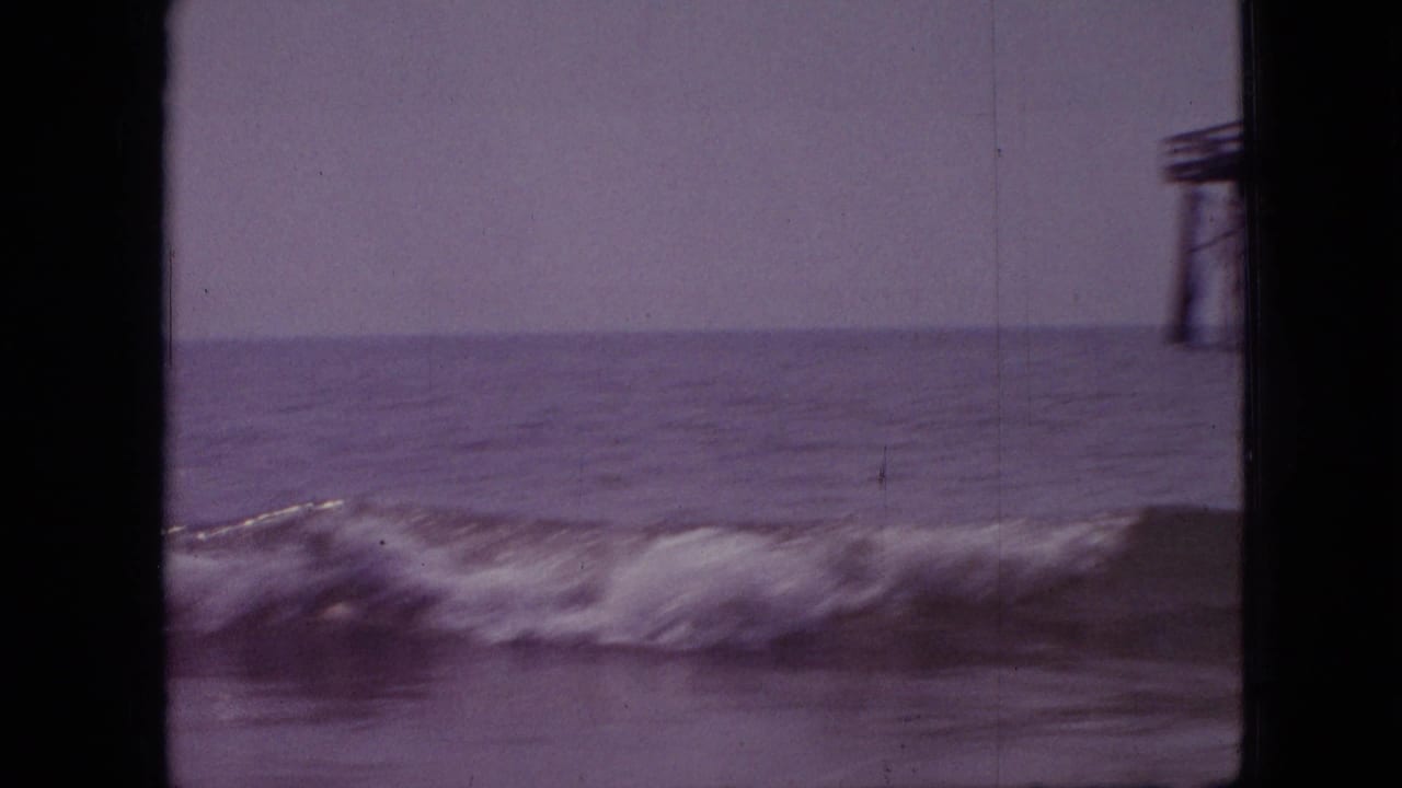 A wave breaks across the waterline while the edge of a pier structure appears at right, captured with soft 8mm grain and a purple color cast.