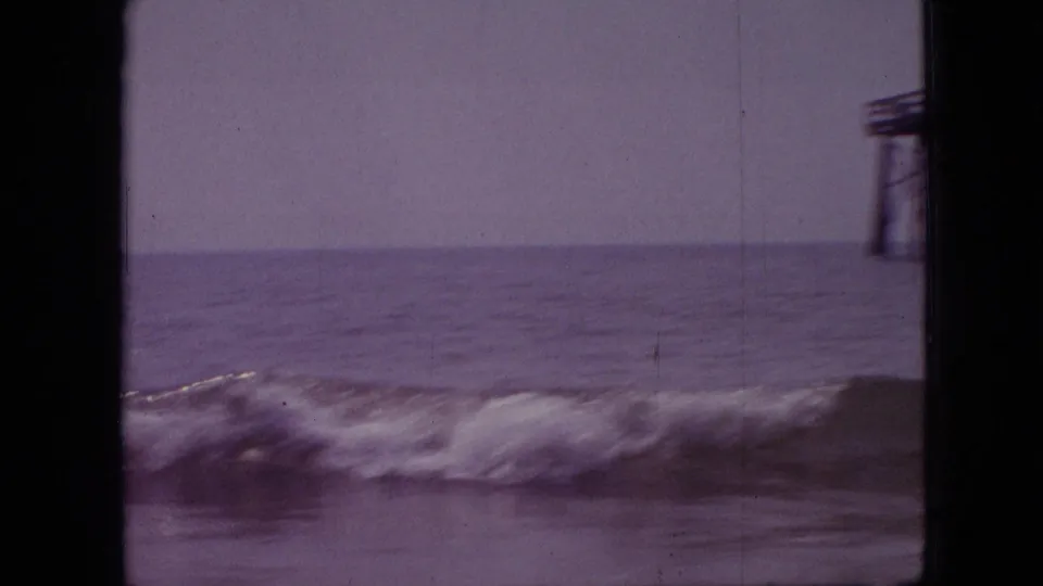 A wave breaks across the waterline while the edge of a pier structure appears at right, captured with soft 8mm grain and a purple color cast.
