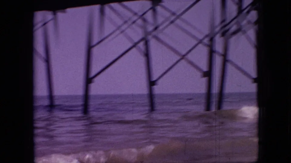 Under-pier supports and diagonal bracing fill the frame above shallow water, with aged 8mm grain, soft focus, and purple color shift.