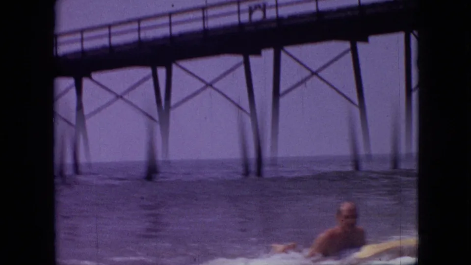 1968 8mm home-movie frame, cataloged as Branson, Missouri, showing an adult male surfer on a board near dark pier supports and small breaking waves.