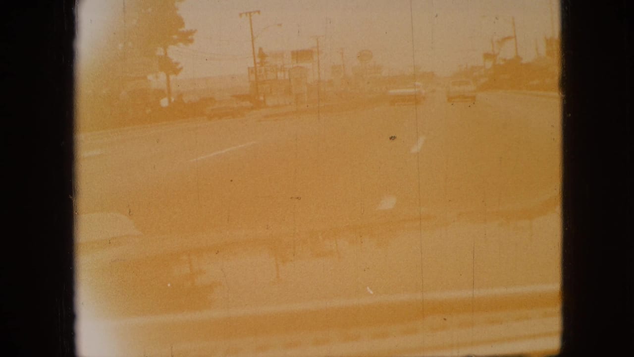 Yellow-tinted 1968 Branson driving view with the car hood at the bottom, roadside buildings, utility poles, and a lightly traveled road ahead.