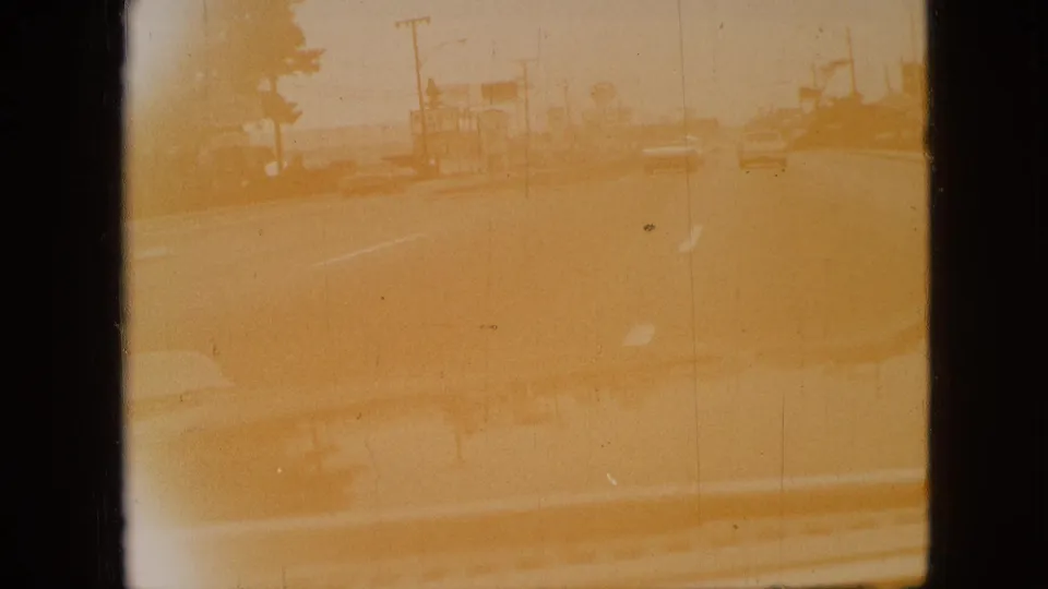 Yellow-tinted 1968 Branson driving view with the car hood at the bottom, roadside buildings, utility poles, and a lightly traveled road ahead.