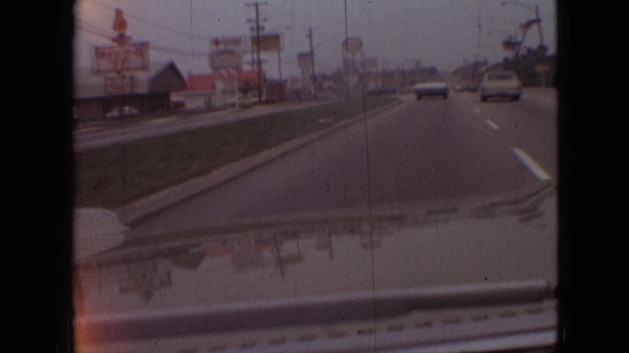 Late-1960s Branson driving frame showing cars ahead on a commercial road, roadside signage, utility poles, a grass strip, and the vehicle hood at the bottom edge.