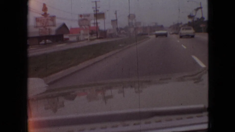 Late-1960s Branson driving frame showing cars ahead on a commercial road, roadside signage, utility poles, a grass strip, and the vehicle hood at the bottom edge.