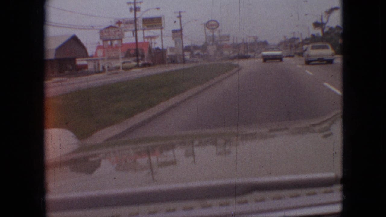 1968 Branson, Missouri 8mm road view from inside a moving car, with roadside business signs, lane markings, distant traffic, and the car hood reflecting the street scene.