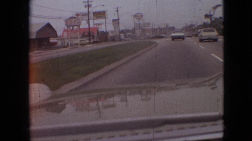 1968 Branson, Missouri 8mm road view from inside a moving car, with roadside business signs, lane markings, distant traffic, and the car hood reflecting the street scene.
