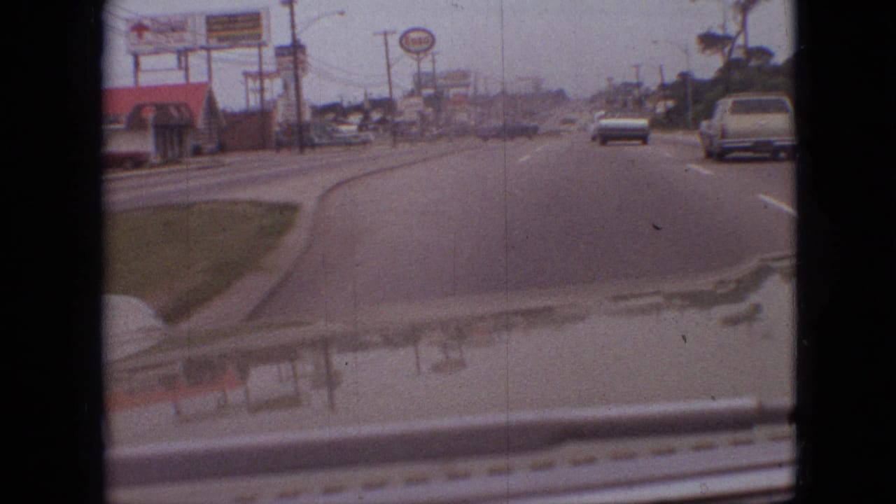 1968 Branson, Missouri roadway seen from a moving car, with a visible Sonic sign, roadside commercial buildings, utility lines, traffic ahead, and reflections across the hood.