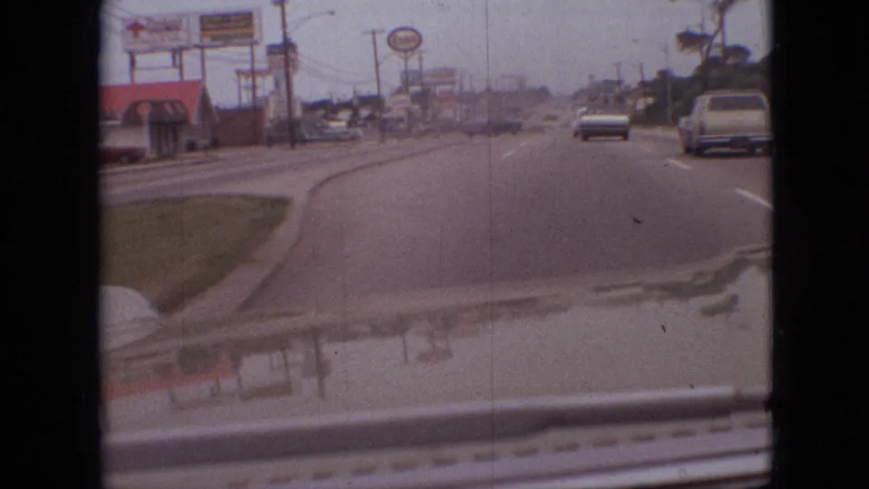 1968 Branson, Missouri roadway seen from a moving car, with a visible Sonic sign, roadside commercial buildings, utility lines, traffic ahead, and reflections across the hood.