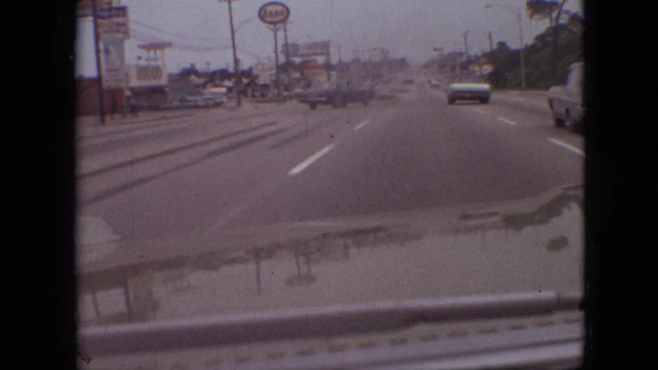 1968 Branson, Missouri moving-car footage showing a commercial roadside, classic traffic, lane markings, a vehicle at the right edge, and the filming car's hood in the foreground.