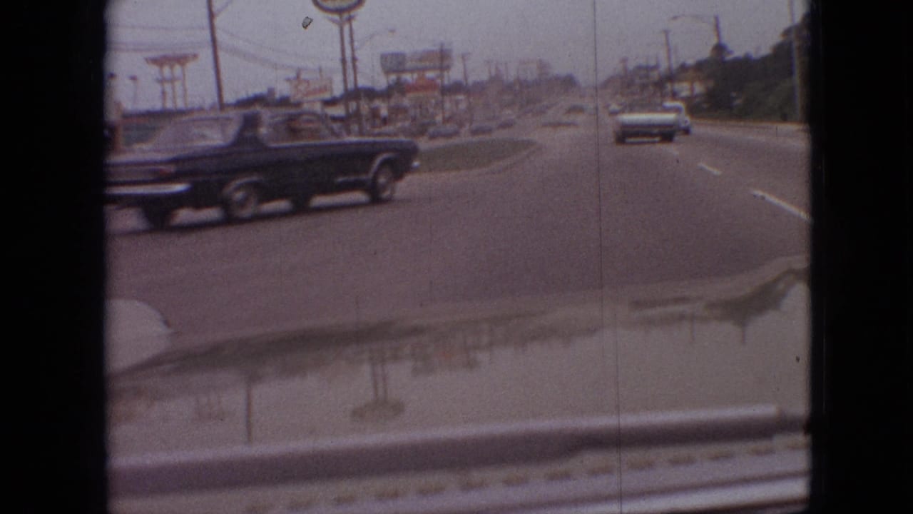 1968 Branson, Missouri road scene from a moving car as a black classic car passes in the foreground, with roadside signage, utility poles, traffic ahead, and hood reflections visible.