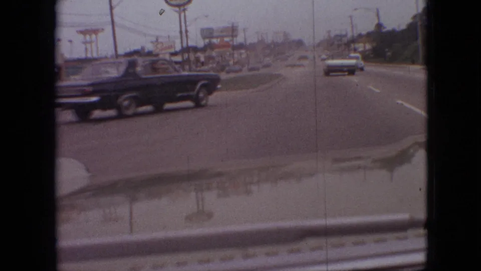1968 Branson, Missouri road scene from a moving car as a black classic car passes in the foreground, with roadside signage, utility poles, traffic ahead, and hood reflections visible.