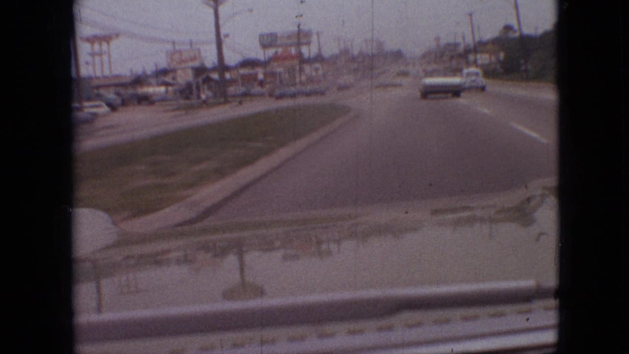 Forward-facing 1968 Branson, Missouri driving view showing a divided street, cars ahead, low commercial buildings, utility poles, and roadside signage under a gray sky.