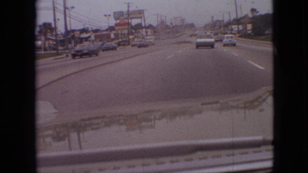 Forward view from a moving vehicle on a Branson commercial roadway in 1968, with period cars, roadside businesses, elevated signs, and a grassy divider visible.