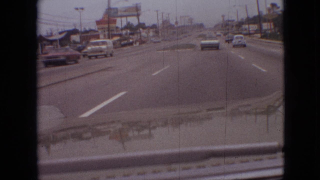 1968 Branson street-driving frame with a dark sedan passing on the left, cars ahead, lane markings, roadside businesses, and tall signs along the corridor.