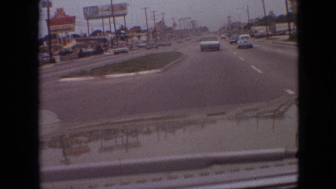 Forward-facing 8mm view of a Branson, Missouri commercial street in 1968, with a grassy median, period cars, roadside signage, low buildings, and windshield reflections.