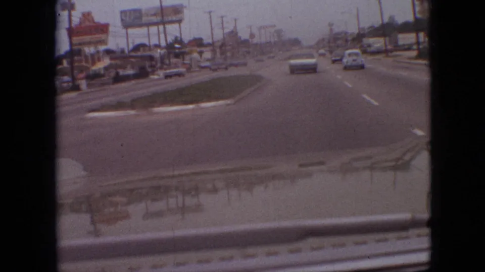 Forward-facing 8mm view of a Branson, Missouri commercial street in 1968, with a grassy median, period cars, roadside signage, low buildings, and windshield reflections.