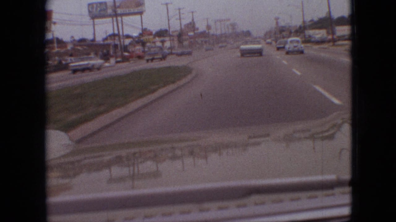 1968 Branson, Missouri road scene filmed from inside a moving vehicle, showing light traffic, billboards, utility poles, and the dashboard edge in the foreground.
