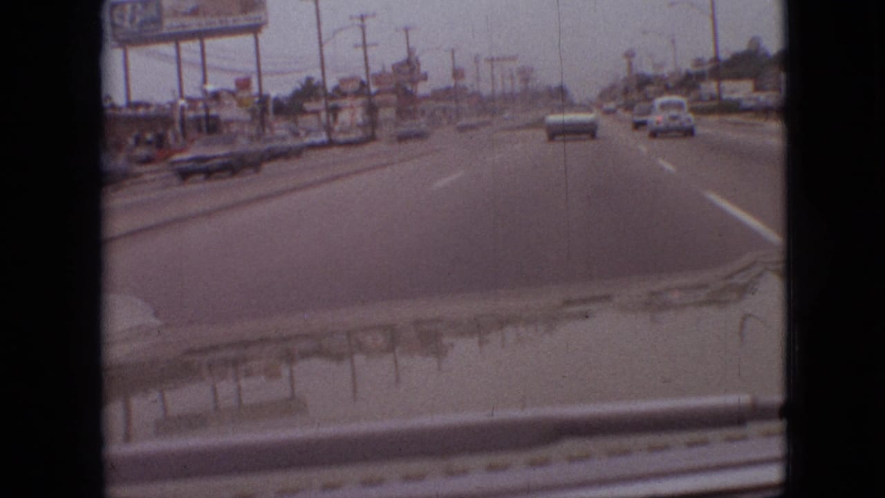 Moving-vehicle view in Branson, Missouri, 1968, with a white sedan ahead on a broad road bordered by signs, poles, and commercial roadside structures.
