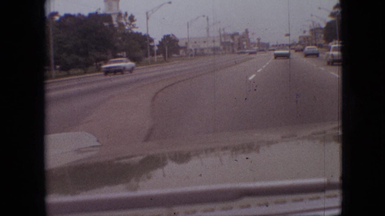 Late-1960s Branson, Missouri road footage from a moving car, with light traffic ahead, a gentle roadway curve, roadside trees, and visible film gate edges.