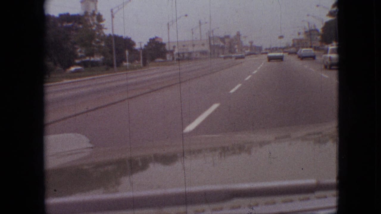First-person 8mm view from a moving car on a multi-lane road in Branson, Missouri, with sedans ahead, streetlights, and low roadside buildings visible under an overcast sky.