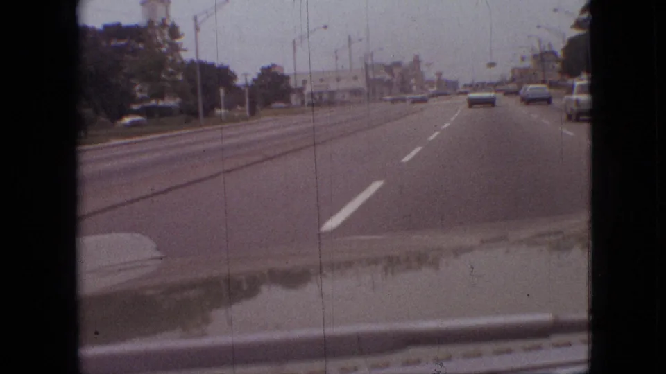 First-person 8mm view from a moving car on a multi-lane road in Branson, Missouri, with sedans ahead, streetlights, and low roadside buildings visible under an overcast sky.