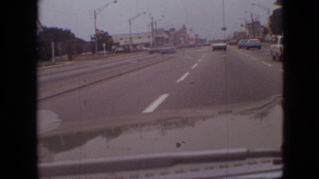 Roadway seen through a windshield with several cars, lane markings, and roadside structures.