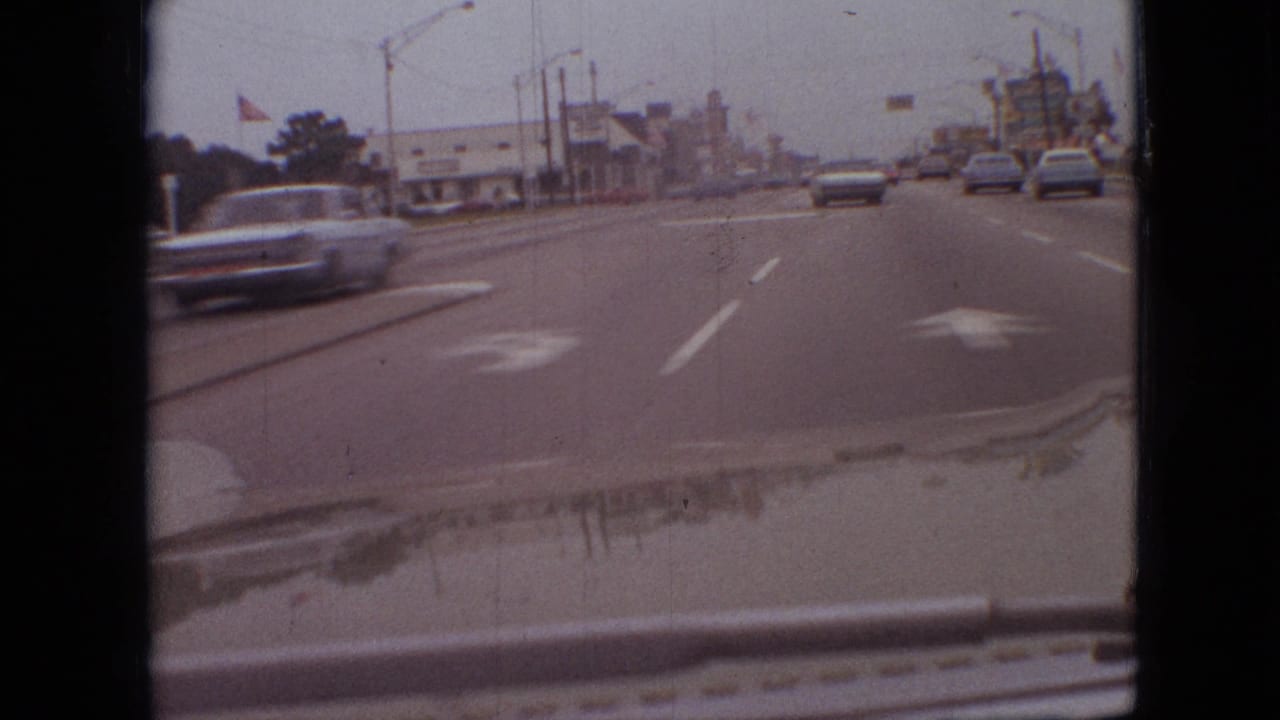 Windshield view of traffic in Branson, Missouri, with a car passing at left, sedans ahead, a roadside flag, streetlights, and commercial buildings along the road.