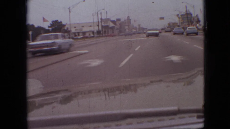 Windshield view of traffic in Branson, Missouri, with a car passing at left, sedans ahead, a roadside flag, streetlights, and commercial buildings along the road.