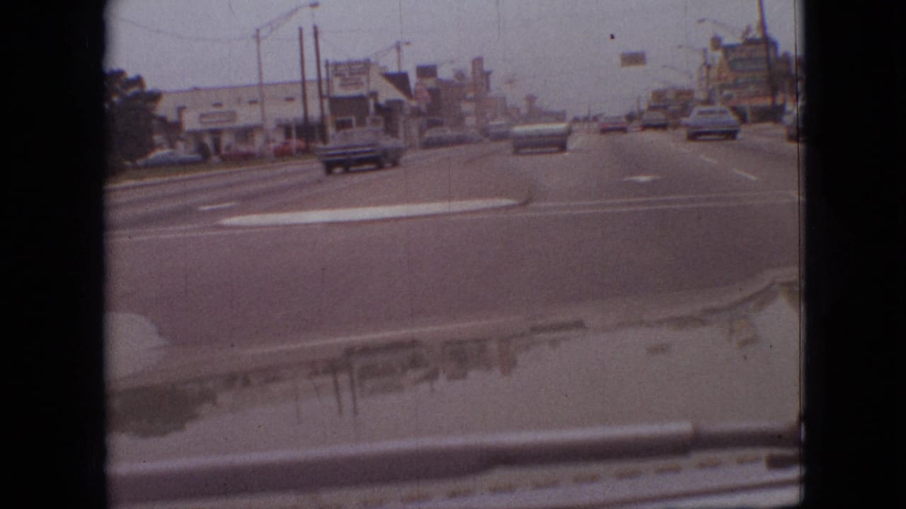 First-person driving view in Branson, Missouri, showing cars ahead on a broad 1968 roadway, low commercial buildings, overhead streetlights, and visible 8mm gate edges.